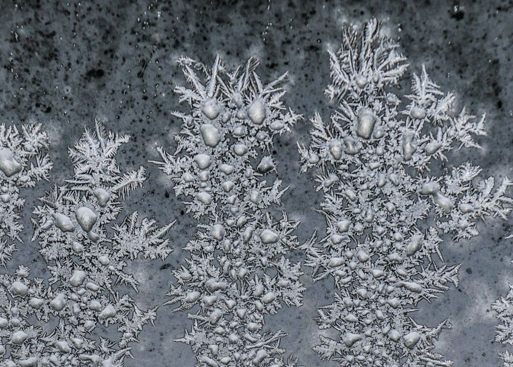 Synthesis Peptides - Close-up of intricate frost patterns and ice crystals on a window, forming dense clusters and delicate, spiky shapes—resembling the complex structures seen in research peptides—set against a dark, mottled background.