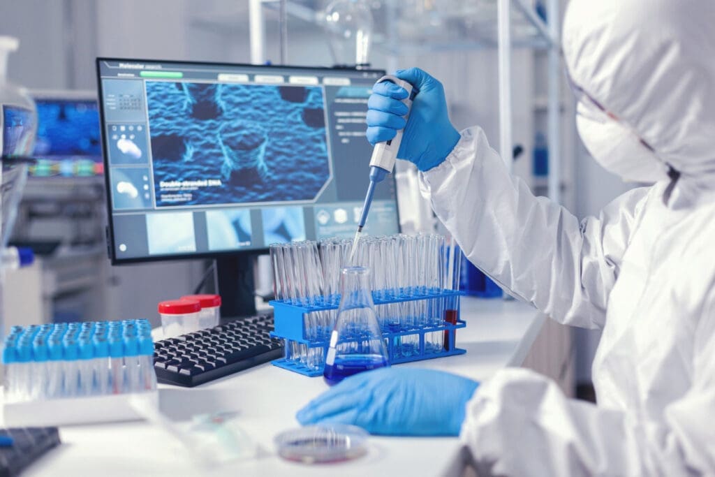Synthesis Peptides - A scientist in protective gear uses a pipette to transfer research peptides into test tubes in a laboratory, with a computer displaying scientific data in the background.